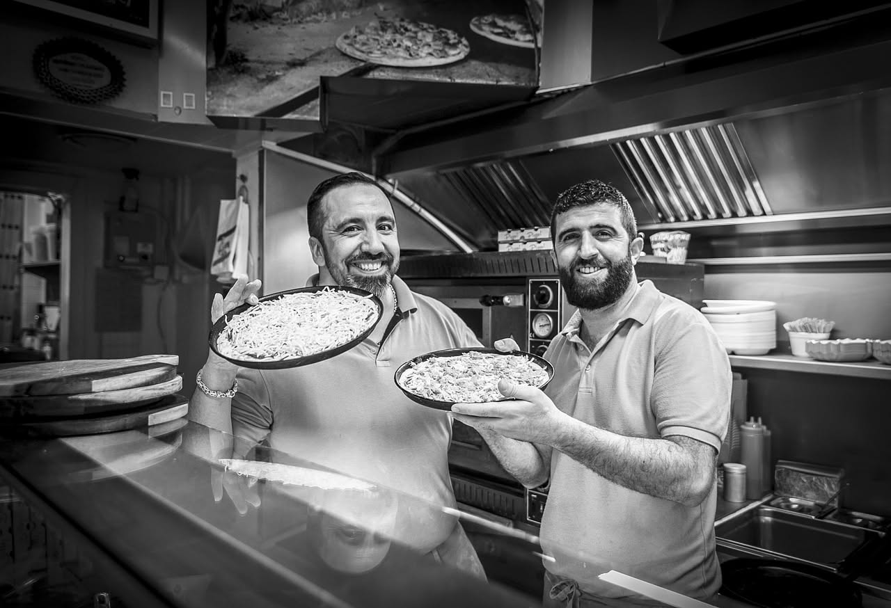 Giovane apprendista in grembiule bianco lavora accanto a un maestro pizzaiolo davanti a un forno a legna in una pizzeria tradizionale napoletana, con le mani che modellan l'impasto