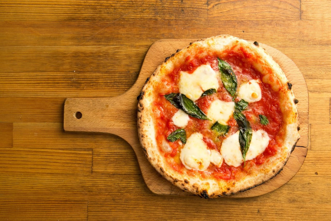 Pizzaiolo che inforna una pizza napoletana nel forno a legna con fiamme visibili sullo sfondo, temperatura manifesta intorno ai 430 gradi