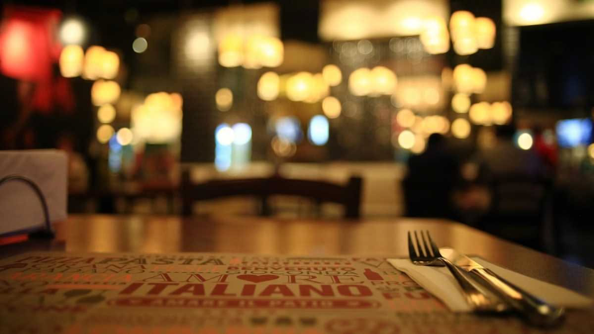 Interno di una pizzeria napoletana con pizzaiolo davanti al forno a legna, tavoli con clienti in sottofondo, banco di servizio e casse