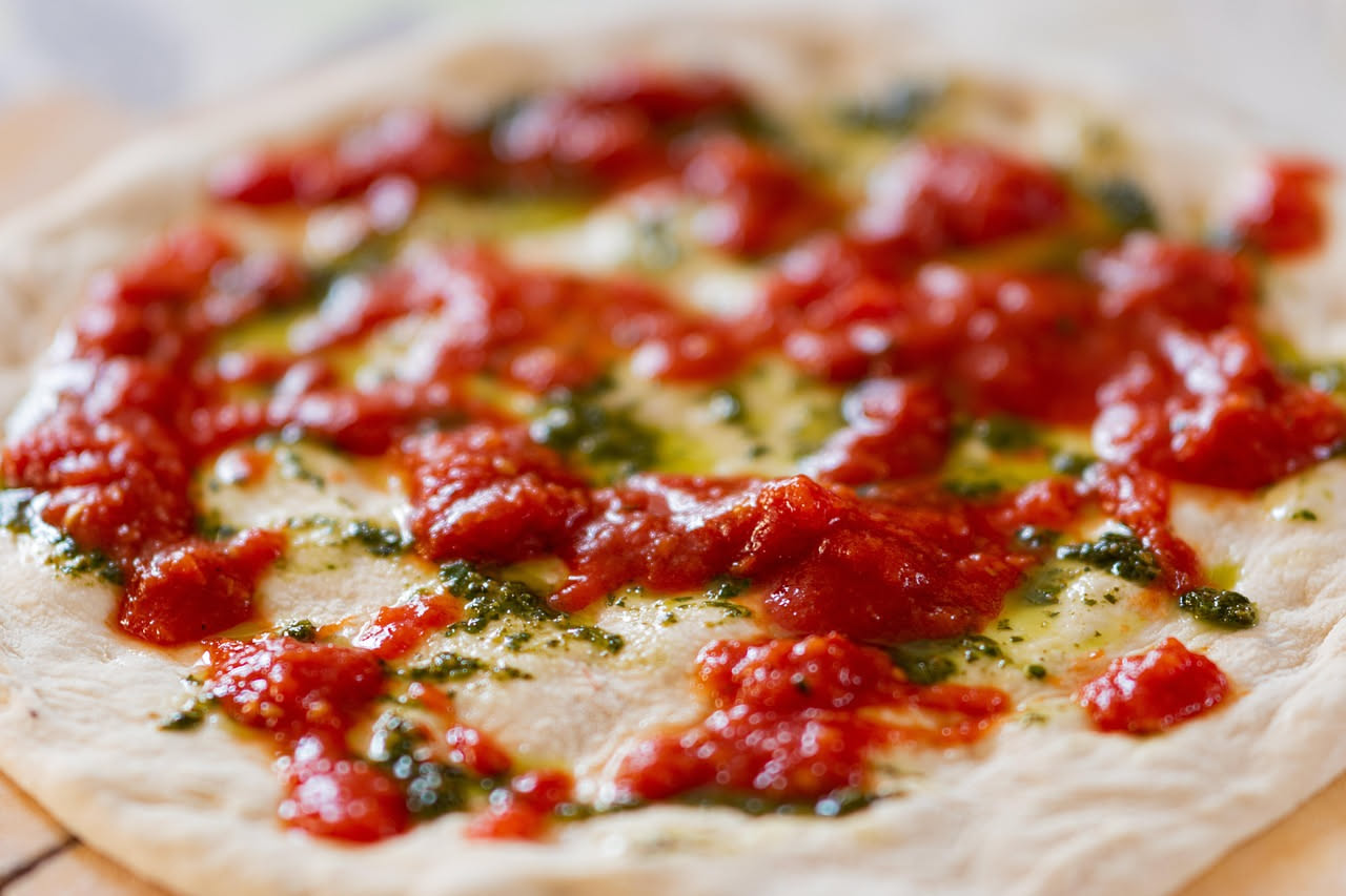Tre bottiglie di vetro scuro contenenti rispettivamente olio extravergine d'oliva verde smeraldo, olio vergine d'oliva giallo dorato e olio di semi trasparente; davanti a una pala di pizza in legno su