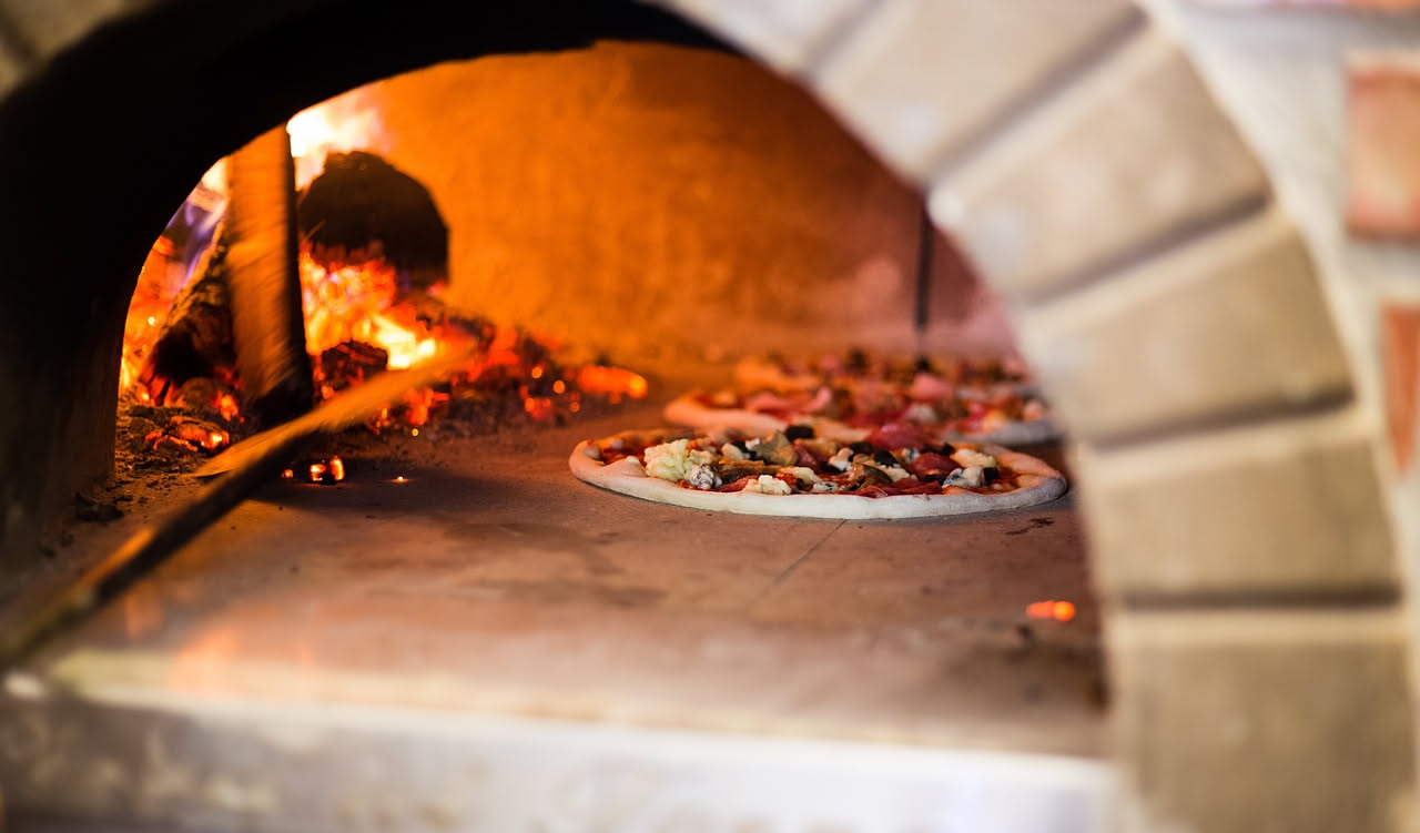 Tre forni a confronto: un forno a legna tradizionale in muratura con bocca semicircolare e legna accatastata, un forno a gas in acciaio con bruciatori visibili, e un forno elettrico compatto moderno c