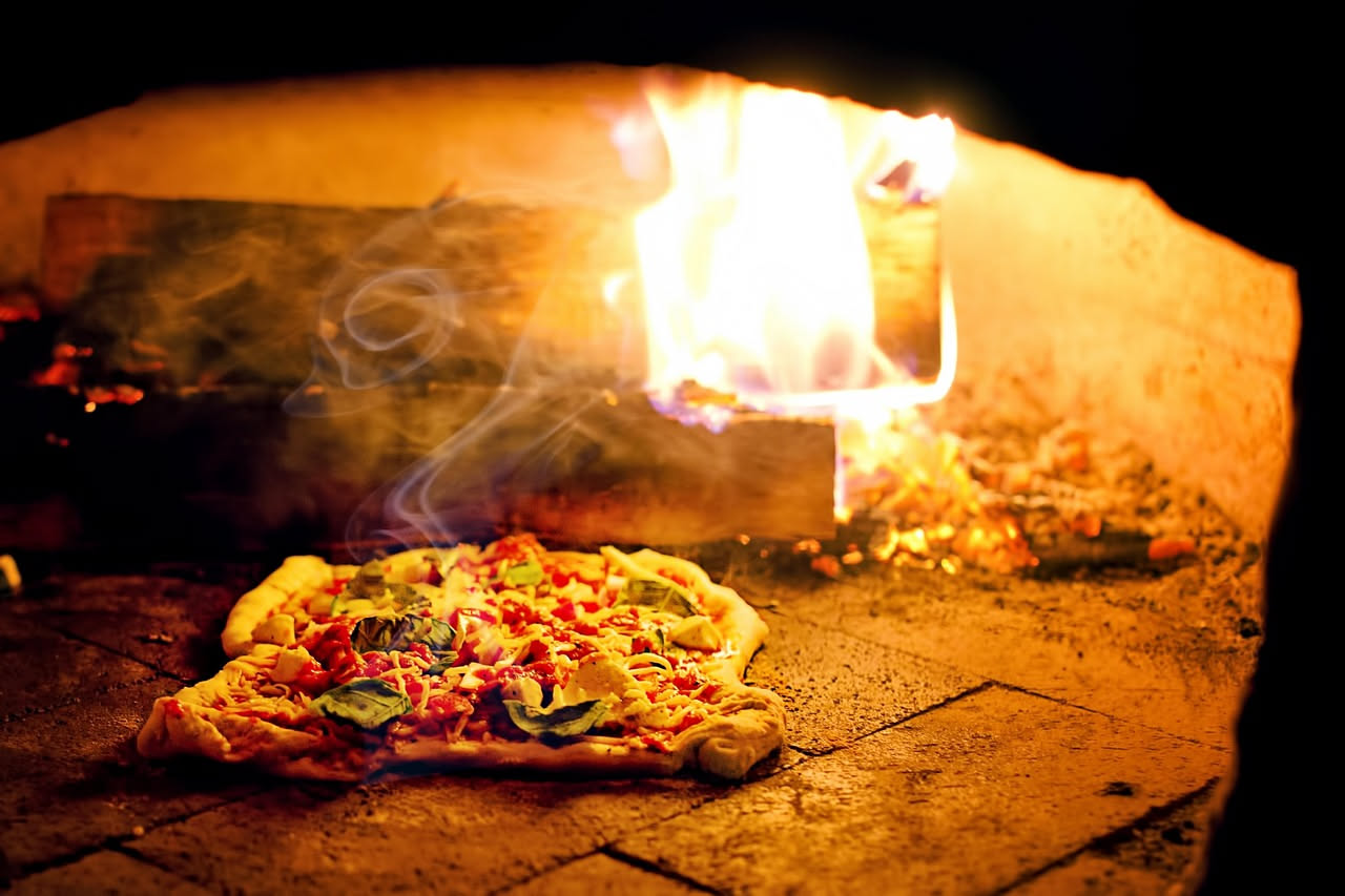 Teglia in terracotta di colore rosso bruno con pizza napoletana dentro, appena sfornata, con cornicione rilevato e mozzarella fusa, ripresa dal lato su sfondo bianco.