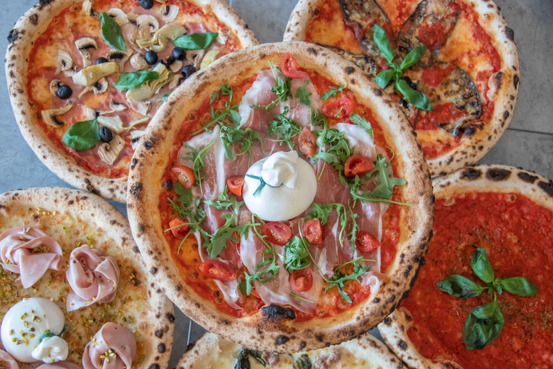 Una pizza al taglio con base dorata appena uscita dal forno, mortadella slicciata appena posata sopra ancora calda, foglie di rucola verde, coltello che taglia la fetta, tavolo in marmo bianco