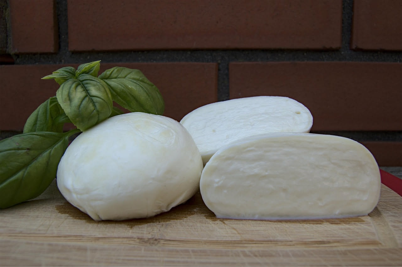 Mozzarella di bufala bianca e cremosa accanto a fiordilatte, su un piatto di legno con basilico fresco e acque di conservazione