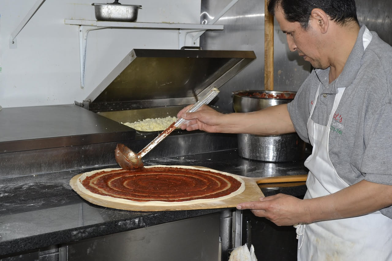 Pizzaiolo con grembiule bianco e farina sulle mani intento a stendere un impasto di pasta cruda su banco di lavoro in legno, con forno a legna sullo sfondo