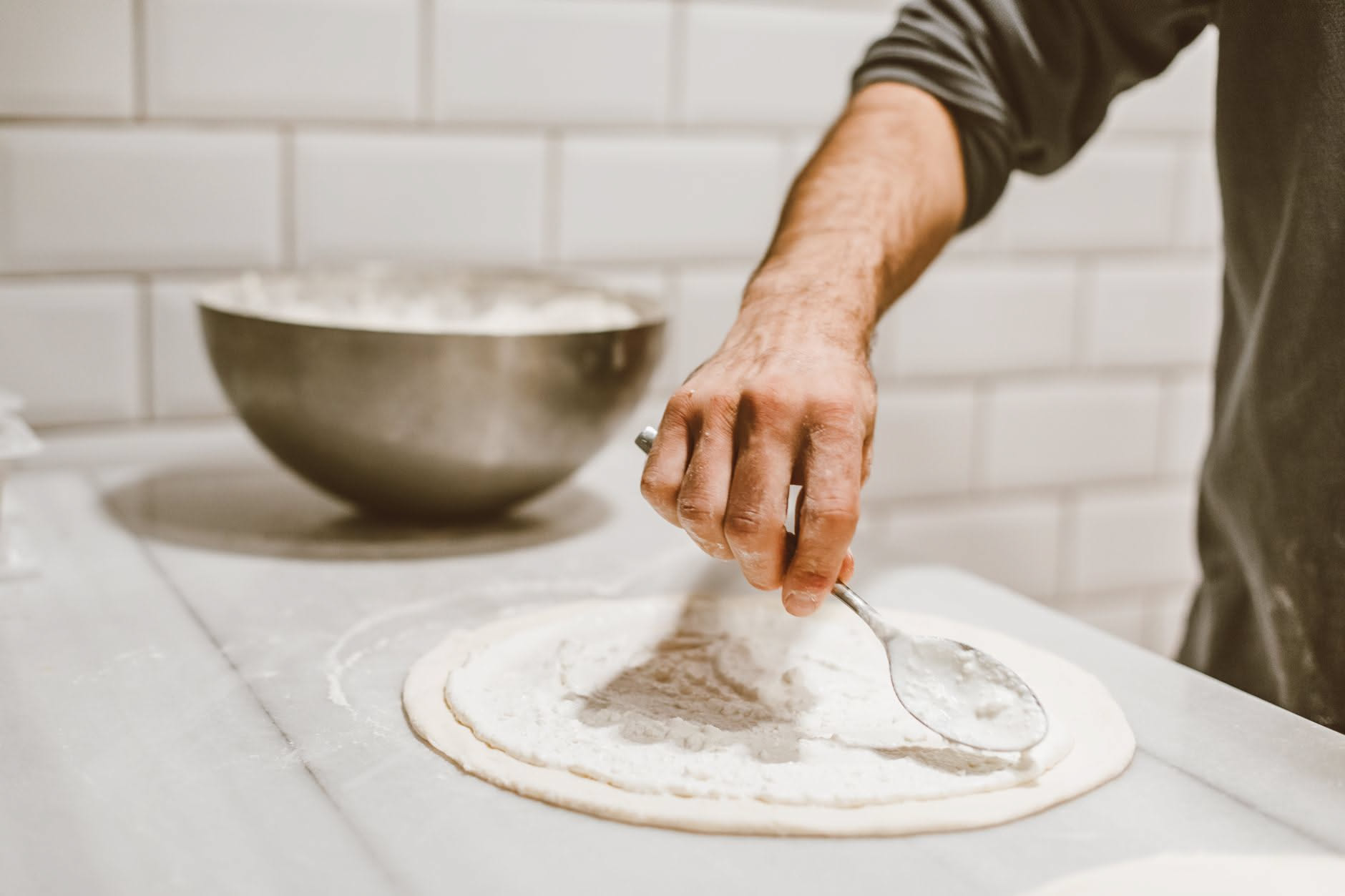 Piano di marmo bianco con impasto di pizza in tre stadi di lievitazione, piccole ciotole di pomodoro e mozzarella, orologio da parete e teglia in acciaio
